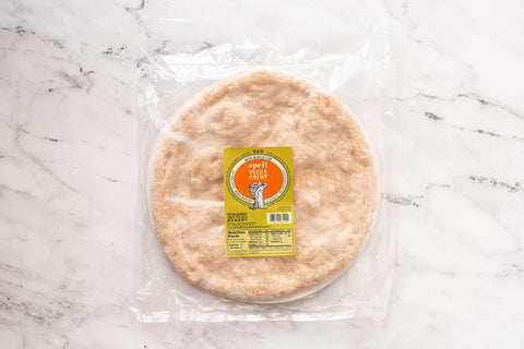 A packaged round spelt pizza crust with a golden-brown, slightly textured surface, photographed from directly above on a white marble surface with gray veining.