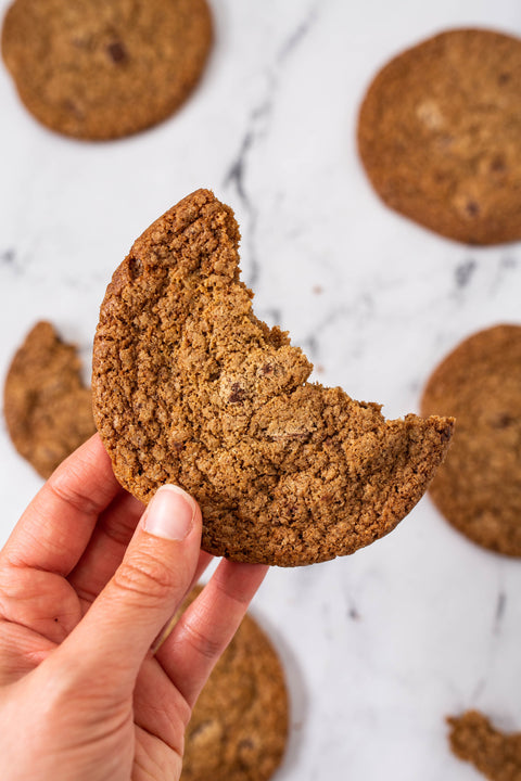 A hand holding a broken half of a thick chocolate chip cookie, with several whole cookies scattered on a white marble surface.