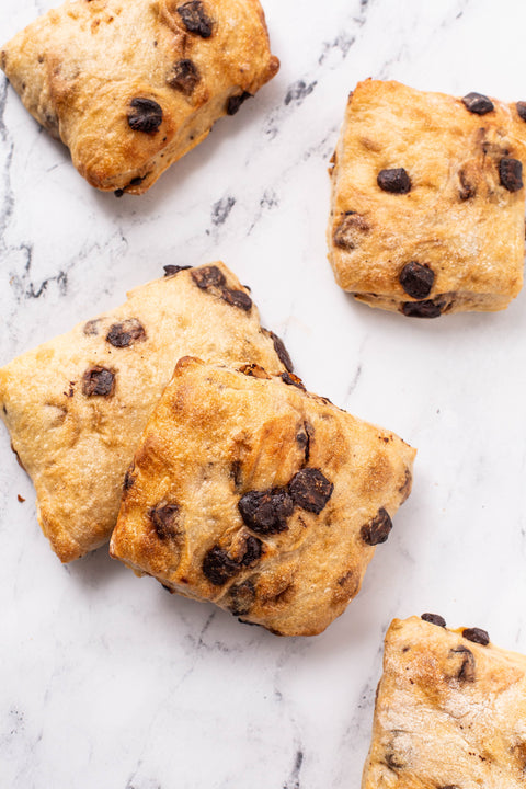 Four baked chocolate chip mini ciabatta with golden-brown crusts and dark chocolate chips, arranged on a white marble surface.