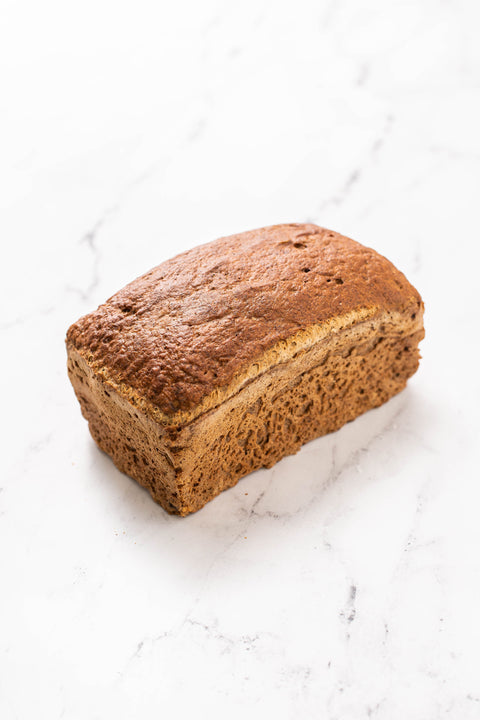 A single loaf of brown bread, Gluten Free Wholegrain Bread, photographed on a white marble surface.
