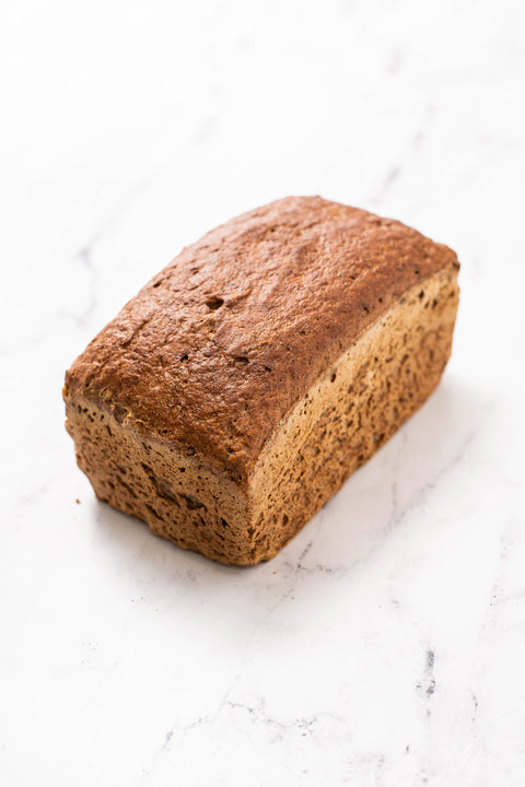 A single loaf of brown bread, Gluten Free Wholegrain Bread, photographed on a white marble surface.