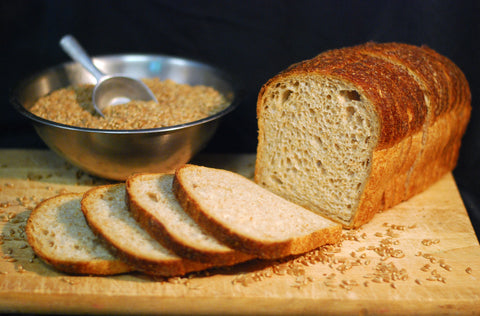 Loaf of peasant french bread with slices on a wooden board next to a bowl of seeds.
