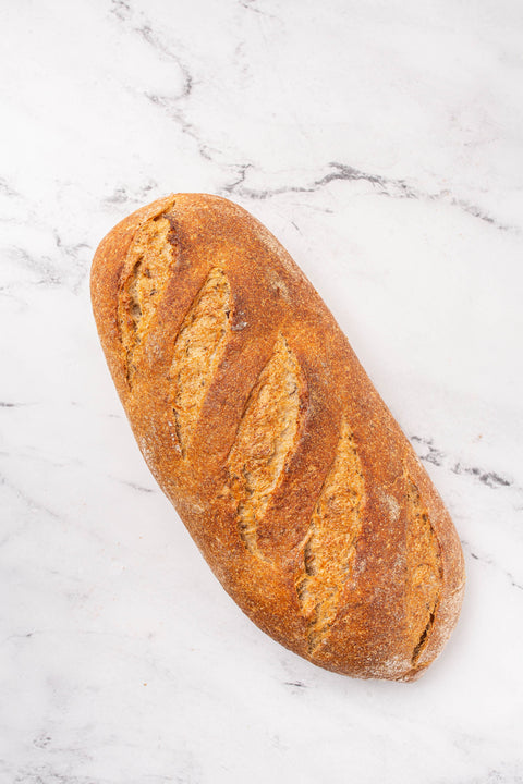 A single rustic artisinal seeded rye loaf of bread with a golden-brown crust and scored slashes, placed diagonally on a white marble surface.