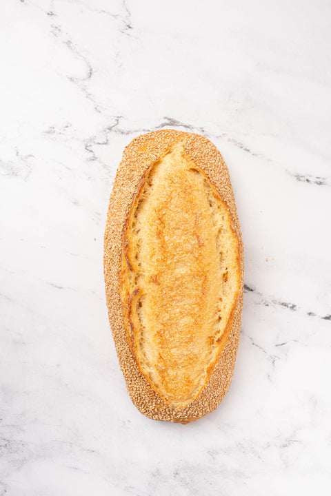 An artisanal baguette with a golden-brown crust and a sesame seed border, presented on a white marble surface.